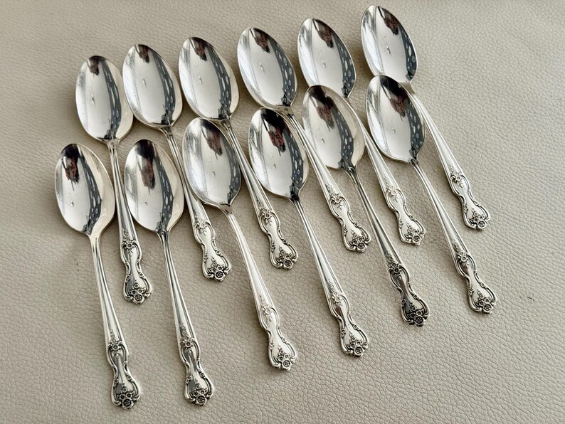 May include: A collection of twelve silver spoons. Each spoon has a rounded bowl and a decorative handle with floral detailing. The spoons are arranged in a fan-like pattern, reflecting light. A classic set for serving.