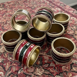 May include: A set of brass napkin rings with red, green, and black enamel stripes. The rings have a gold-coloured interior and are accented with a beaded trim. The napkin rings are arranged on a patterned red and brown surface.