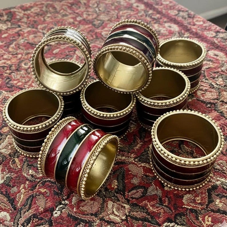 May include: A set of brass napkin rings with red, green, and black enamel stripes. The rings have a gold-coloured interior and are accented with a beaded trim. The napkin rings are arranged on a patterned red and brown surface.