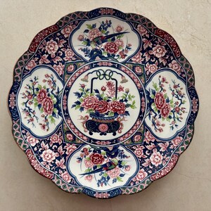 Pu&ograve; includere: Un piatto rotondo in ceramica con bordo smerlato caratterizzato da un medaglione centrale con un cesto di fiori circondato da quattro pannelli contenenti uccelli e fiori. Il design utilizza una tavolozza di blu scuro, bianco, rosa e verde.