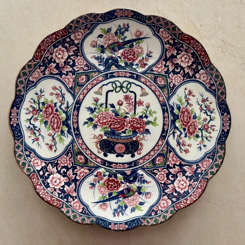Pu&ograve; includere: Un piatto rotondo in ceramica con bordo smerlato caratterizzato da un medaglione centrale con un cesto di fiori circondato da quattro pannelli contenenti uccelli e fiori. Il design utilizza una tavolozza di blu scuro, bianco, rosa e verde.