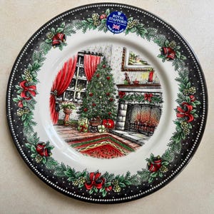 以下が含まれることがあります： ロイヤルスタッフォードの装飾用プレート。クリスマスのシーンが描かれています。黒い縁には白いドットとお祝いの緑が施されています。中央には、装飾されたクリスマスツリー、暖炉、赤いカーテンが描かれています。