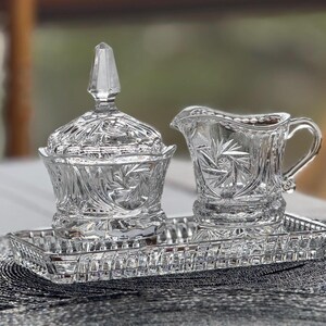 May include: A clear pressed glass set consisting of a lidded sugar bowl, a creamer pitcher, and a rectangular serving tray. Each piece features intricate starburst and geometric patterns with scalloped edges and a pointed finial on the lid.