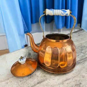 May include: A copper tea kettle with a fluted body, a curved spout, and a brass handle featuring a white ceramic grip with blue floral patterns. The matching copper lid has a small white ceramic knob with a blue floral design on top.