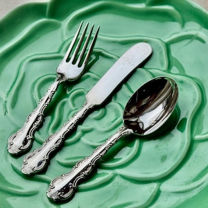 May include: A silver fork, knife, and spoon set on a green, floral-shaped plate. The cutlery has ornate, detailed handles. The plate is a light green colour, and the silverware is reflective.