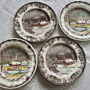May include: Four brown and white ceramic dinner plates with a winter scene featuring a red church, trees, and snow. The plates have a brown rim and a white center with a brown and green floral pattern.