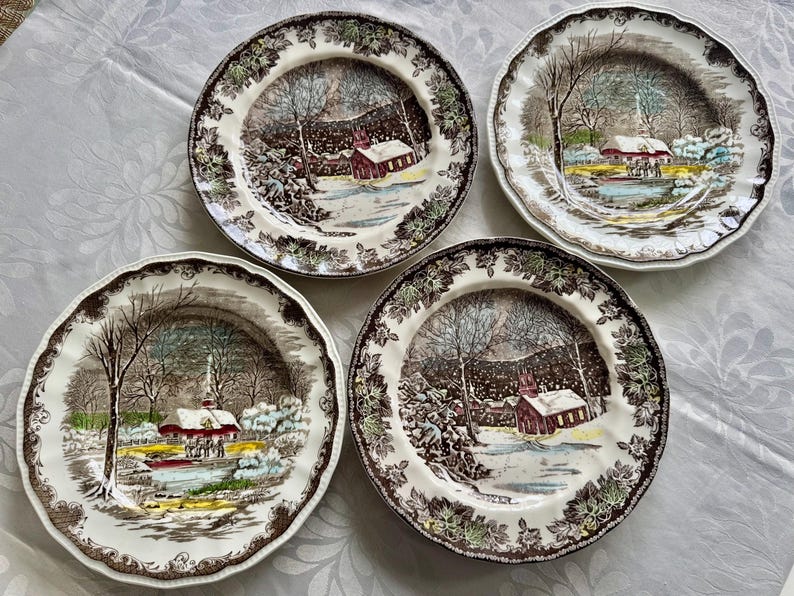 May include: Four brown and white ceramic dinner plates with a winter scene featuring a red church, trees, and snow. The plates have a brown rim and a white center with a brown and green floral pattern.