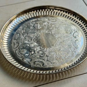 Puede incluir: Una bandeja redonda plateada con un dise&ntilde;o floral en relieve. La bandeja tiene un borde decorativo y acanalado. La bandeja es probablemente de metal y es adecuada para servir alimentos o exhibir objetos decorativos.