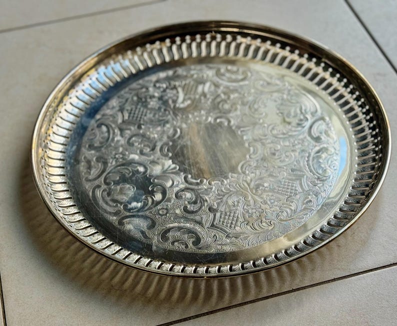 Puede incluir: Una bandeja redonda plateada con un dise&ntilde;o floral en relieve. La bandeja tiene un borde decorativo y acanalado. La bandeja es probablemente de metal y es adecuada para servir alimentos o exhibir objetos decorativos.