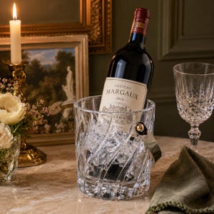 May include: A clear, heavy-cut crystal wine bucket with a swirling pattern and a metallic handle, filled with ice cubes. A bottle of Chateau Margaux 2016 red wine is placed inside the bucket, with the label clearly visible against the ice.