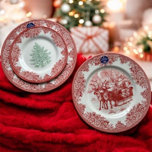 May include: Set of Christmas-themed plates with red floral borders. One plate features a green Christmas tree, while another depicts Santa Claus in a sleigh pulled by reindeer. The plates are on a red surface, with a blurred Christmas tree in the background.