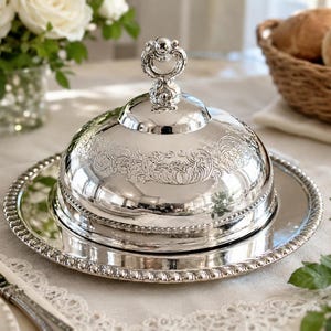 May include: A silver butter dish with a domed lid and ornate detailing. The dish sits on a matching round tray with a beaded edge. The lid features a decorative handle. The butter dish is on a white tablecloth, with a basket of bread in the background.
