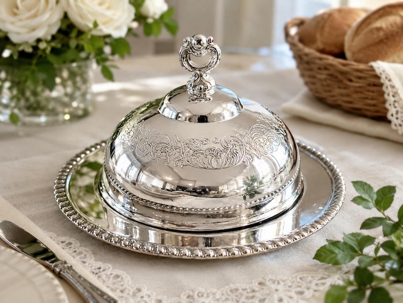 May include: A silver butter dish with a domed lid and ornate detailing. The dish sits on a matching round tray with a beaded edge. The lid features a decorative handle. The butter dish is on a white tablecloth, with a basket of bread in the background.