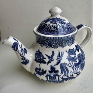 K&ouml;nnte beinhalten: Eine wei&szlig;e Teekanne mit einem blauen und wei&szlig;en Weidenmuster. Die Teekanne hat einen runden K&ouml;rper und einen Deckel mit einem Knopf oben. Die Teekanne ist mit einem traditionellen chinesischen Design aus V&ouml;geln, Blumen und einer Br&uuml;cke verziert.