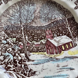 May include: A vintage ceramic plate with a winter scene featuring a church, houses, and trees covered in snow. The plate has a brown and white color scheme and a delicate floral border.