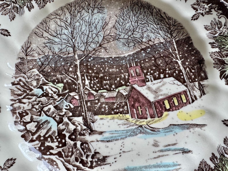 May include: A vintage ceramic plate with a winter scene featuring a church, houses, and trees covered in snow. The plate has a brown and white color scheme and a delicate floral border.