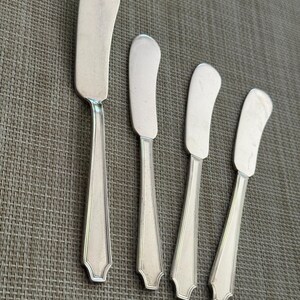 May include: Four silver butter spreaders with curved blades and decorative handles. The blades are a bright silver, and the handles feature a detailed design. The spreaders are arranged on a textured surface.