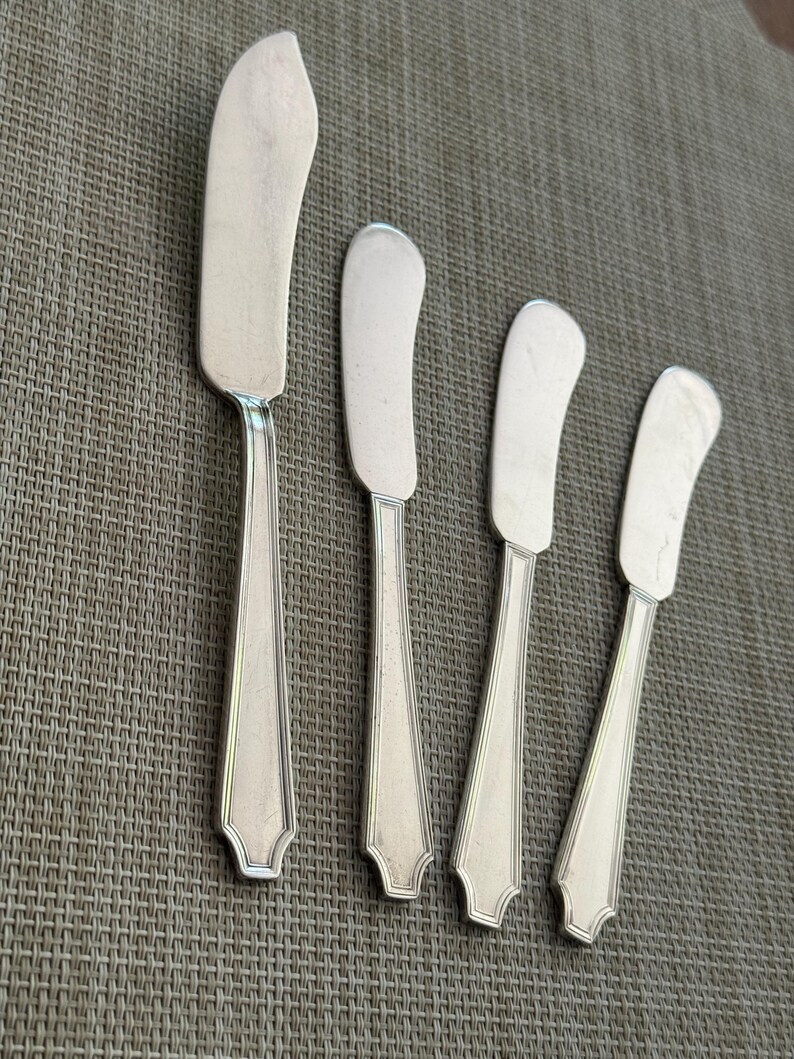 May include: Four silver butter spreaders with curved blades and decorative handles. The blades are a bright silver, and the handles feature a detailed design. The spreaders are arranged on a textured surface.