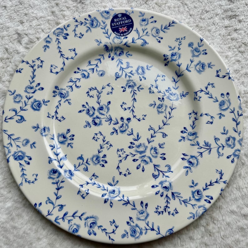 May include: A round ceramic dinner plate with a cream-colored base and a repeating blue floral pattern of roses and vines. A circular blue sticker at the top reads Royal Stafford Est 1845 Made in England with a small Union Jack flag icon included.