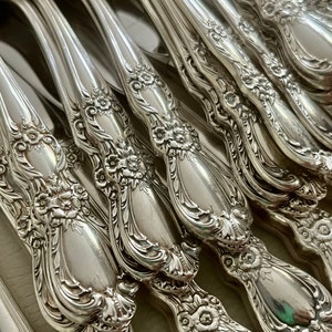 May include: Close-up of a collection of ornate silver-colored flatware. The handles feature a floral design with intricate detailing. The silverware includes knives, forks, and spoons, all with a polished finish.