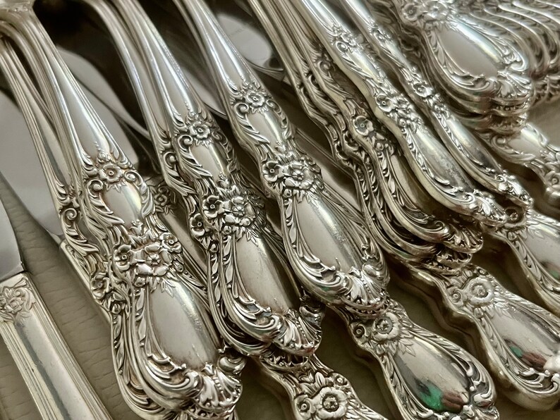May include: Close-up of a collection of ornate silver-colored flatware. The handles feature a floral design with intricate detailing. The silverware includes knives, forks, and spoons, all with a polished finish.