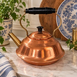 May include: A polished copper tea kettle with a wide, rounded base and a tapered spout. It features a black wooden handle attached to brass-colored metal supports and a matching black knob on the removable lid, creating a classic kitchen aesthetic.
