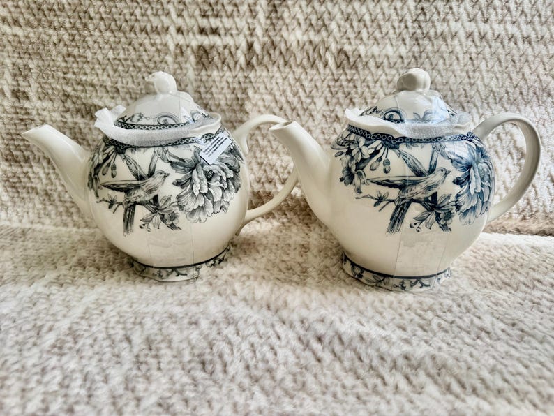 K&ouml;nnte beinhalten: Zwei wei&szlig;e Keramik-Teekannen mit blauem Blumen- und Vogelmuster. Jede Teekanne hat einen Deckel, einen Ausguss und einen Griff. Die Deckel haben einen dekorativen Rand. Die Teekannen stehen auf einer strukturierten Oberfl&auml;che.