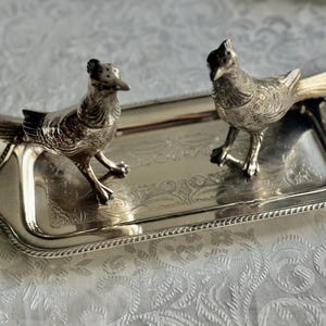 May include: A silver-colored serving tray with two decorative bird figurines. The tray has a detailed, ornate design and a beaded edge. The birds are detailed with textured feathers and stand on the tray.