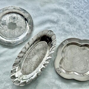 May include: Three antique silver serving trays. The trays have ornate floral and scrollwork patterns. One is round, one is oval, and one is a quatrefoil shape. The trays are displayed on a light blue patterned tablecloth.