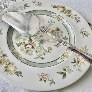 K&ouml;nnte beinhalten: Ein silberner Tortenheber liegt auf einem wei&szlig;en Porzellanteller mit floralem Muster. Der Teller hat einen gr&uuml;nen Rand und Blumen in Gelb, Blau und Rot. Der Heber hat einen dekorativen Griff.