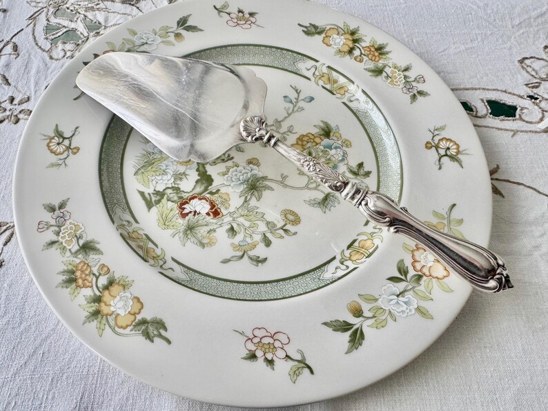K&ouml;nnte beinhalten: Ein silberner Tortenheber liegt auf einem wei&szlig;en Porzellanteller mit floralem Muster. Der Teller hat einen gr&uuml;nen Rand und Blumen in Gelb, Blau und Rot. Der Heber hat einen dekorativen Griff.