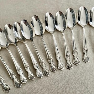 May include: A collection of silver spoons with ornate floral detailing on the handles. The spoons are arranged in a diagonal pattern, reflecting light and creating a shiny appearance. The spoons are likely for serving.