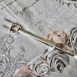 May include: A silver-toned cake lifter with a two-pronged fork at one end and a diamond-shaped handle at the other. The lifter is set against a floral patterned background with roses in shades of gray and beige.