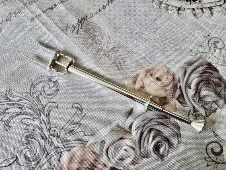 May include: A silver-toned cake lifter with a two-pronged fork at one end and a diamond-shaped handle at the other. The lifter is set against a floral patterned background with roses in shades of gray and beige.