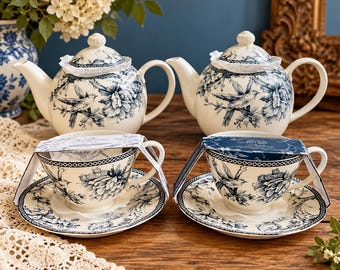May include: Two cream-colored ceramic teapots and two matching teacups with saucers, all featuring a blue floral and bird pattern. Each piece includes a decorative paper band wrapped around the top, showcasing a consistent toile-style design.