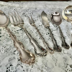 May include: A collection of antique silver-plated flatware, including a spoon, forks, knife, and serving spoons. The silverware has ornate, detailed handles and a reflective surface, arranged on a white lace tablecloth.