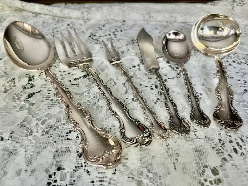 May include: A collection of antique silver-plated flatware, including a spoon, forks, knife, and serving spoons. The silverware has ornate, detailed handles and a reflective surface, arranged on a white lace tablecloth.