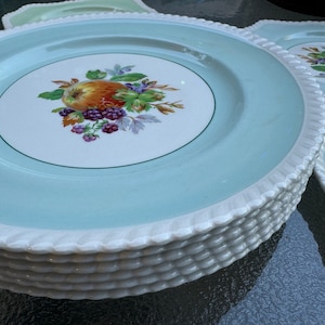 Puede incluir: Una pila de platos de cena de cer&aacute;mica azul claro y blanco con un borde verde y un dise&ntilde;o floral con una manzana roja, moras y hojas verdes.