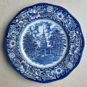 May include: A round ceramic plate featuring a blue and white transferware pattern. The center depicts a historic building with a tall steeple, trees, and figures. The wide border is decorated with a dense arrangement of flowers and leaves.