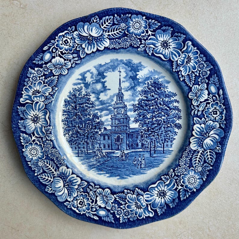 May include: A round ceramic plate featuring a blue and white transferware pattern. The center depicts a historic building with a tall steeple, trees, and figures. The wide border is decorated with a dense arrangement of flowers and leaves.