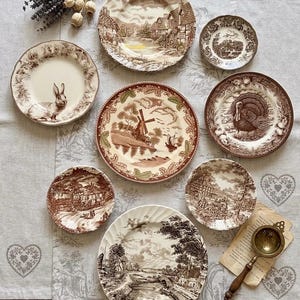 May include: A collection of eight ceramic plates featuring brown transferware patterns, including scenes of windmills, rural landscapes, a turkey, and a rabbit, arranged on a light gray fabric runner with a metal tea strainer nearby.