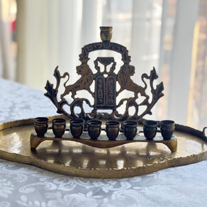 May include: A brass menorah with seven candle holders, set on a hammered brass tray. The menorah features a decorative design with lions and Hebrew text. The tray has handles and a slightly oval shape. The background is blurred.