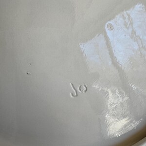 May include: A close-up of a cream-colored ceramic plate. The plate has a glossy finish and the initials "Jo" are stamped on the surface. The plate is slightly off-white.