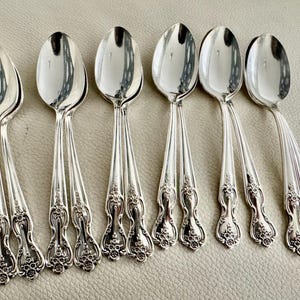May include: Six silver spoons with ornate floral detailing on the handles. The spoons have a reflective, polished surface, and are arranged in a row. The spoons are likely for dining or serving.