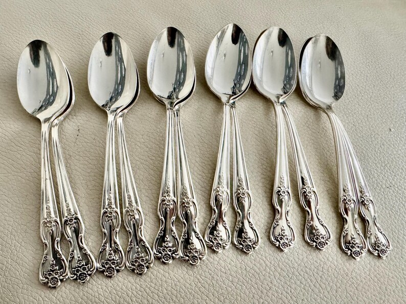 May include: Six silver spoons with ornate floral detailing on the handles. The spoons have a reflective, polished surface, and are arranged in a row. The spoons are likely for dining or serving.