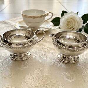 Puede incluir: Un juego de azucarero y lechera plateados con asas ornamentadas y una banda decorativa alrededor del centro. Al fondo, una taza de t&eacute; de porcelana blanca con borde dorado sobre un plato a juego, acompa&ntilde;ada por una rosa blanca.