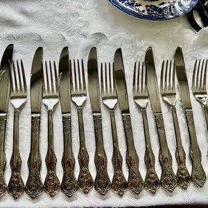 May include: An assortment of antique silverware, including forks and knives, arranged on a white tablecloth. The silverware has ornate handles and a shiny, metallic finish, suggesting a formal dining setting.
