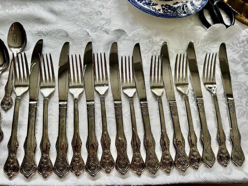 May include: An assortment of antique silverware, including forks and knives, arranged on a white tablecloth. The silverware has ornate handles and a shiny, metallic finish, suggesting a formal dining setting.