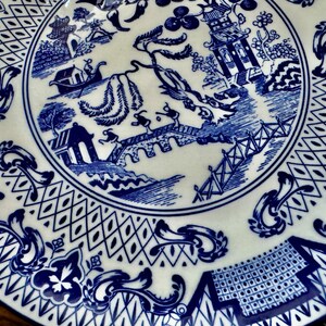 K&ouml;nnte beinhalten: Ein wei&szlig;er Keramikteller mit blauem Willow-Muster. Der Teller zeigt eine zentrale Szene mit einer Br&uuml;cke, Figuren und Geb&auml;uden, umgeben von einem dekorativen Rand. Ein klassisches St&uuml;ck Geschirr.