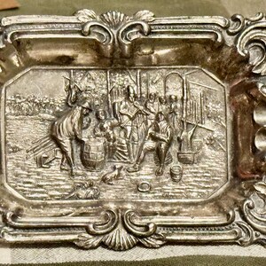May include: A rectangular silver-toned metal ashtray featuring an ornate, raised border with scrollwork and shell motifs. The center depicts a detailed relief scene of several figures gathered around a barrel in an outdoor setting.